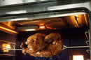 Close-up view inside an oven showing a whole rotisserie chicken being roasted on a spit rod beneath an active infrared heating element, with golden-brown crispy skin illuminated by interior lighting.